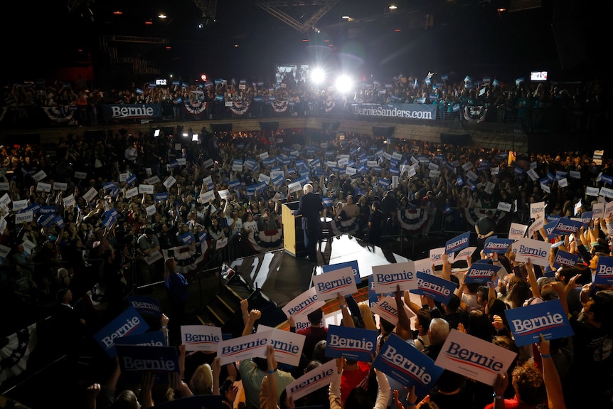 Bernie Sanders stands on stage surrounded by supporters holding signs are a rally.