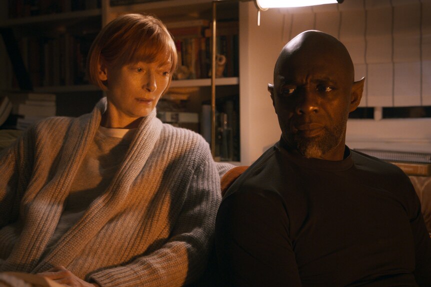 Two middle-aged people - a woman and a Black man - sit on a couch in a dim room, her looking at him