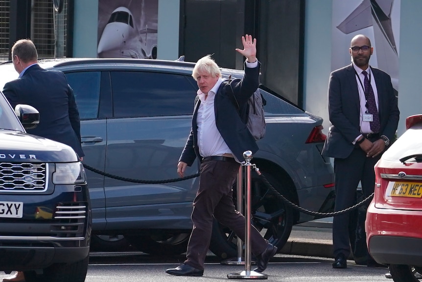 Boris walks in front of a car and waves. Two security guards stand either side of him. 