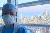 A nurse wearing PPE (a face mask, cap, scrubs and gown) stands in front of a window, the London skyline on a clear sunny day