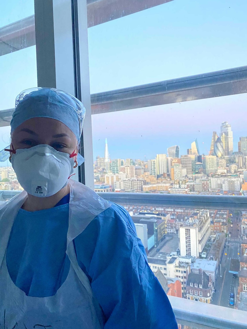 A nurse wearing PPE (a face mask, cap, scrubs and gown) stands in front of a window, the London skyline on a clear sunny day