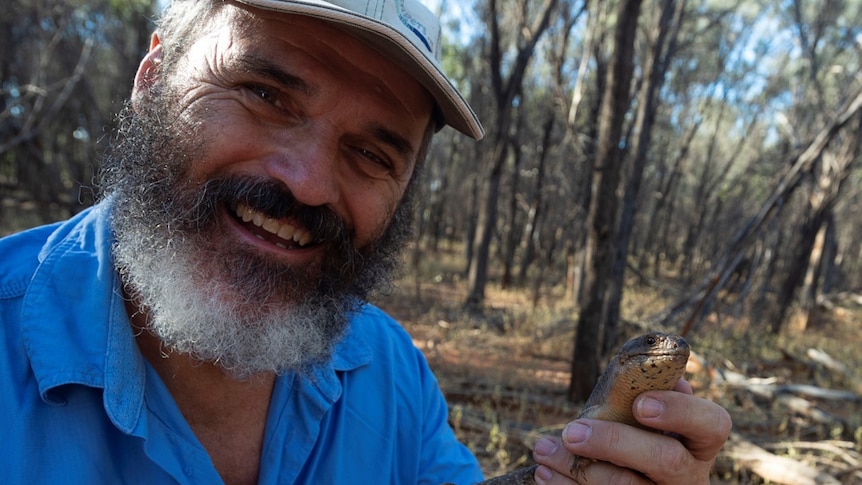 A man in a blue button up shirt and a cap holding a small lizard.