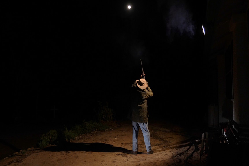 a man shoots his shotgun, pointed towards a pitch-black sky with the moon shining