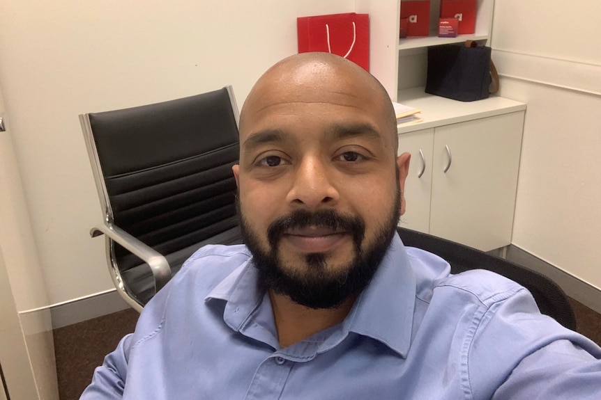 A man with facial hair, wearing a blue business shirt, takes a selfie. 