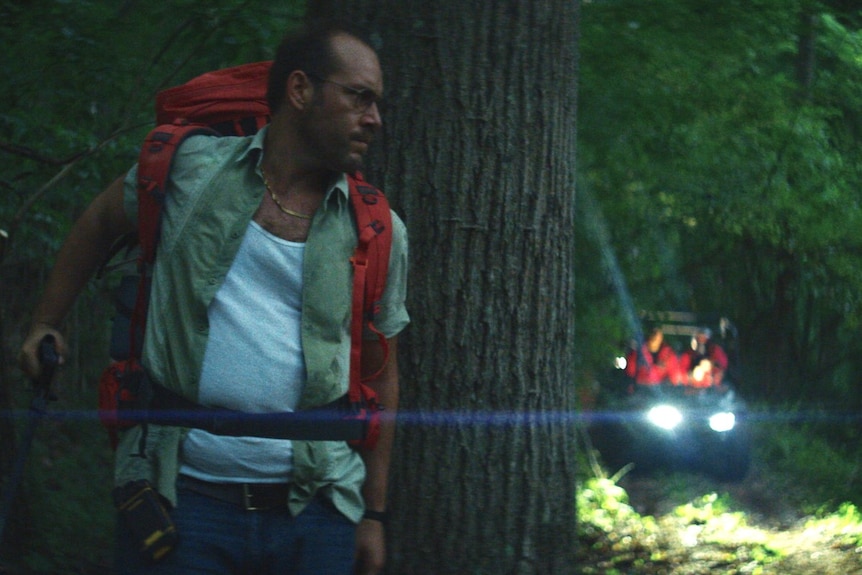 Dean Imperial, a middle aged man with a backpack his back against a tree in a dark night, in the film Lapsis