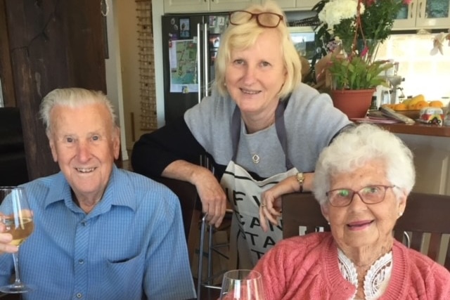 Woman leans on the back of two chairs where her parents are sitting.