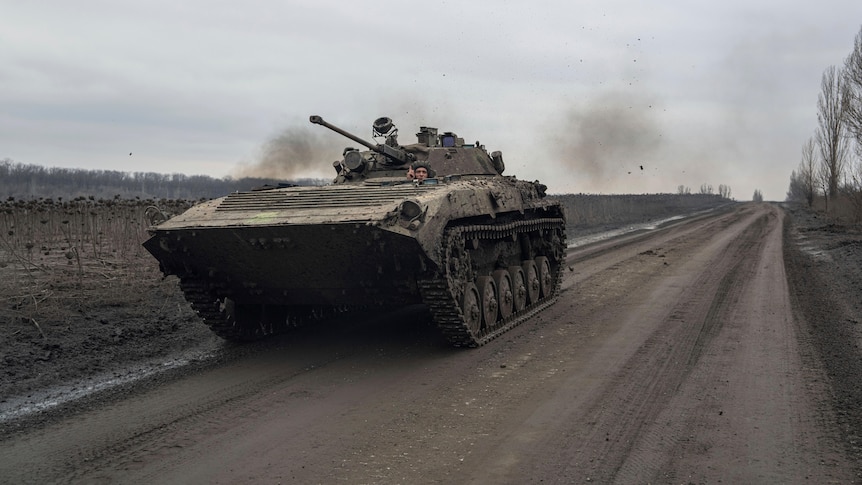 A Ukrainian serviceman drives an armoured personnel carrier along a muddy dirt road past stripped barren fields. 
