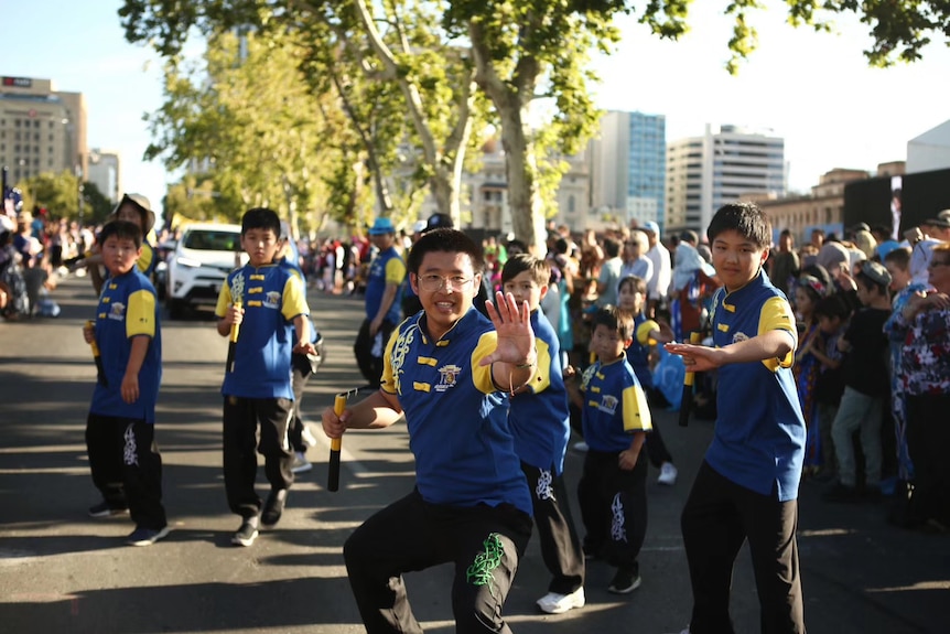 A young and small Asian man in blue and yellow does a kungfu move on a street parade surrounded by smiling fellow students.