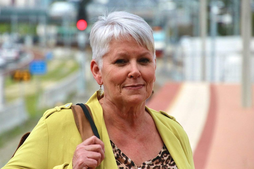 Jill wearing a yellow shirt and holding a bag over her shoulder, standing on a railway platform.