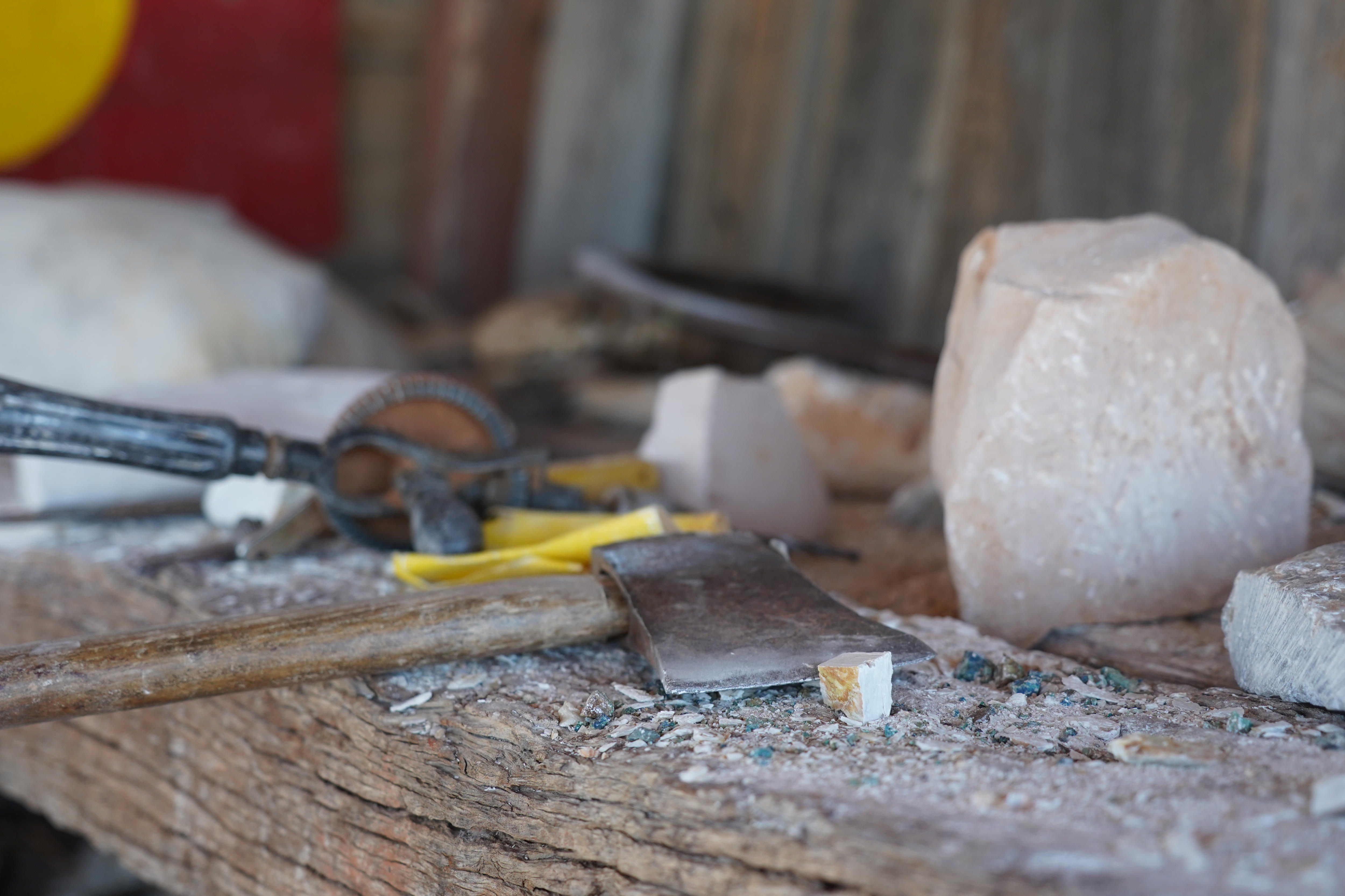 Una vieja hacha pequeña y papel de lija se encuentran sobre un banco de trabajo de madera rodeado de trozos de piedra blanca. 