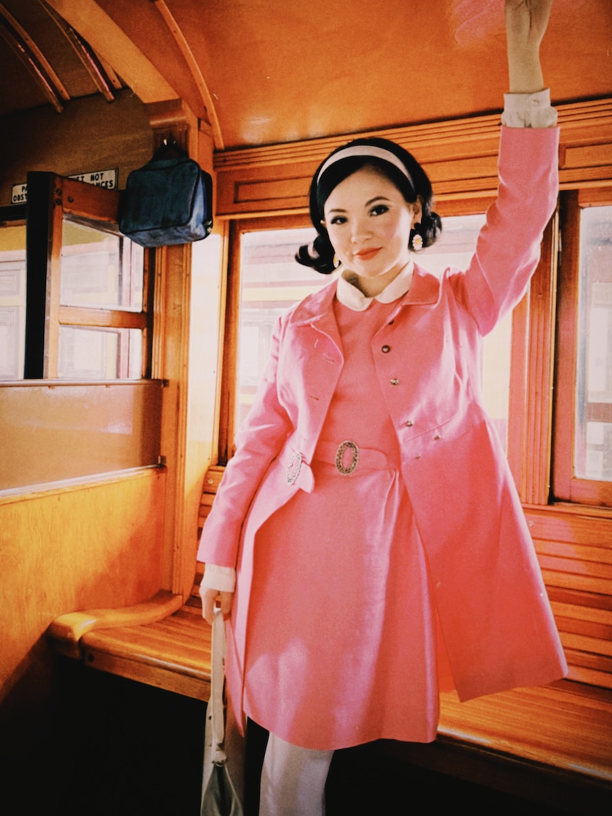 A young woman in an old style pram wearing a light pink dress and coat