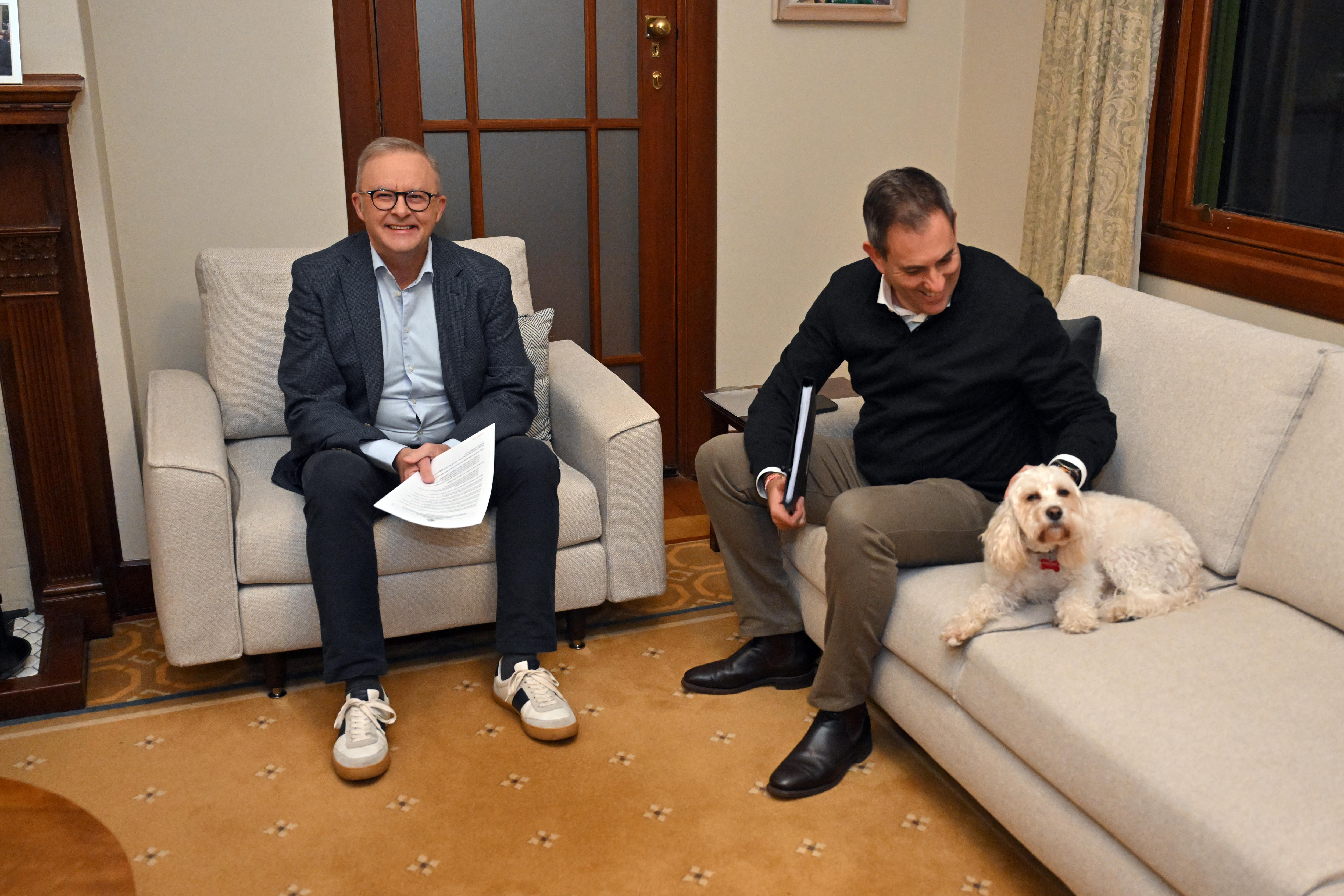 Albanese and Chalmers sit on lounges in a living room smiling, as Chalmers pets a small fluffy white dog.
