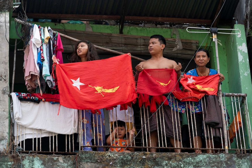 Two women and a shirtless man hold red NLD flags as they stand on a balcony.