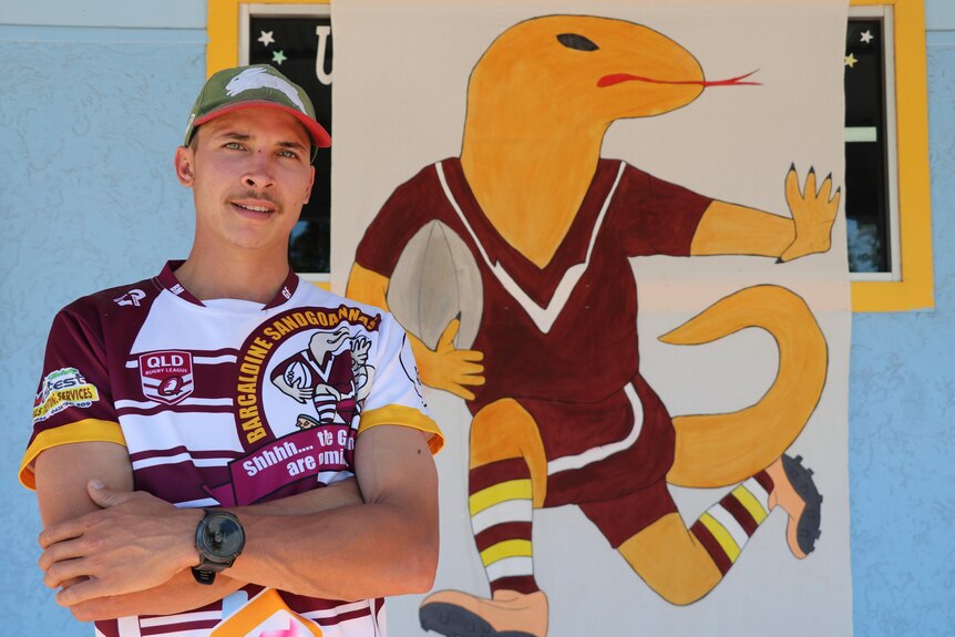 a young man wearing a maroon jersey stands in front of a huge poster with the barcaldine sandgoanna logo on it in his home town