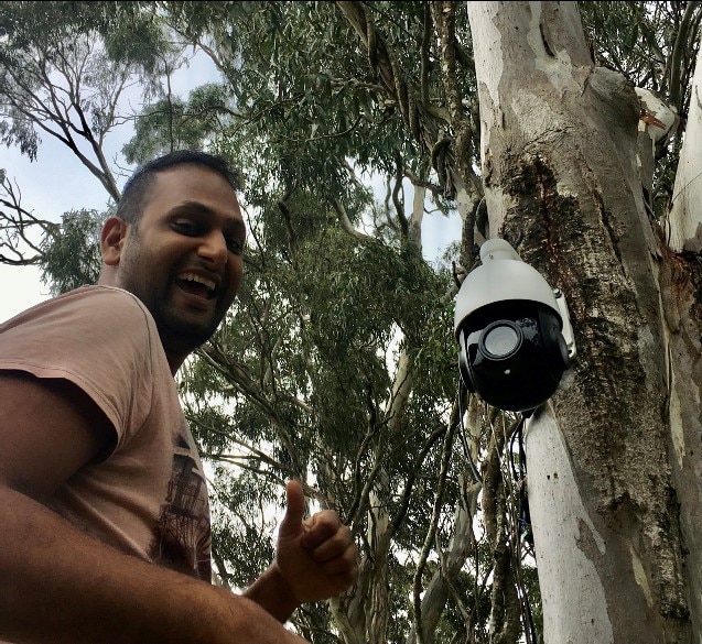 A man smiling with an automated camera attached to a tree. 
