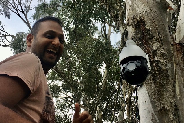 A man smiling with an automated camera attached to a tree. 