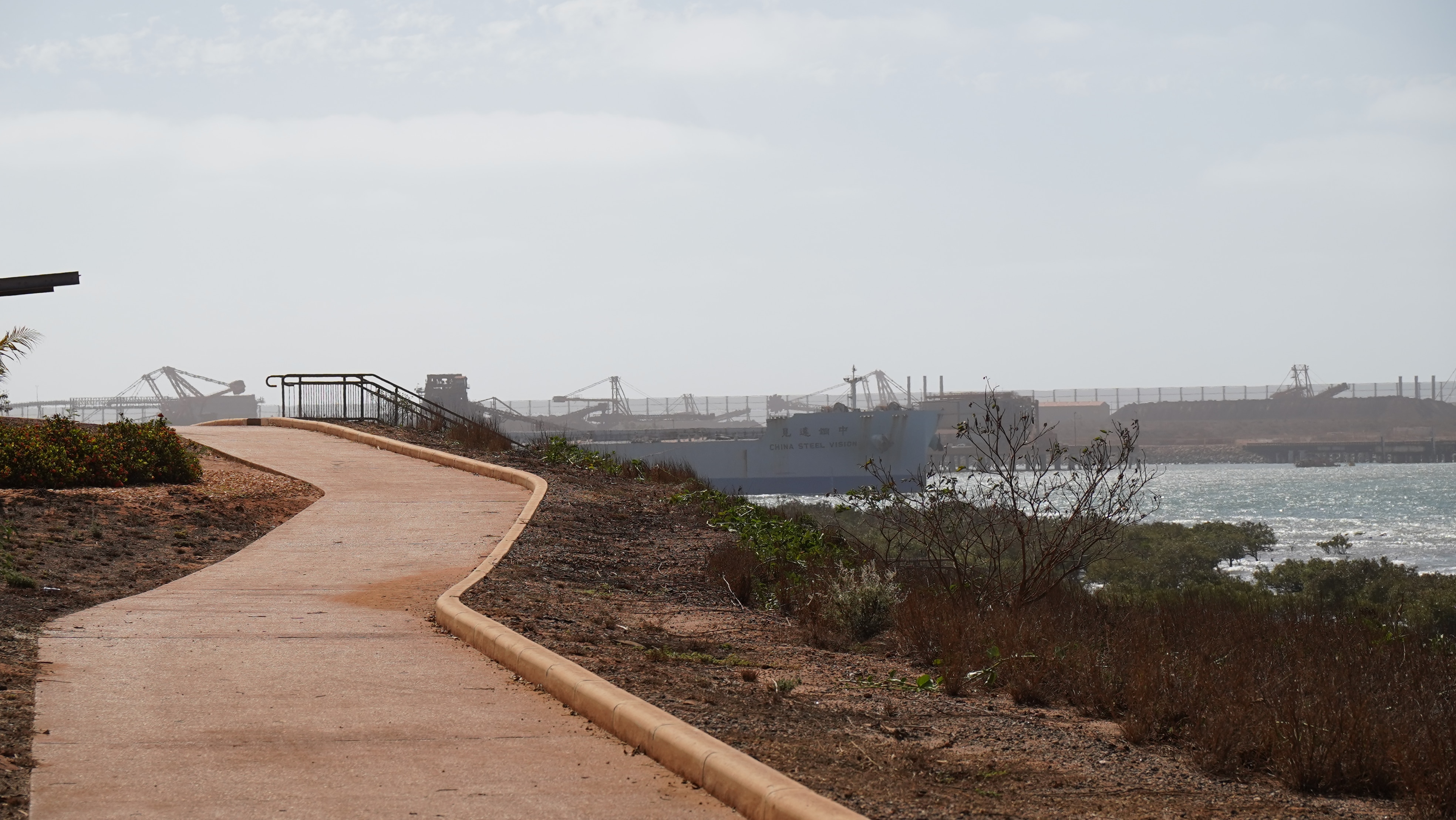 Un sendero cubierto de polvo naranja junto al mar, con grandes barcos industriales al fondo.
