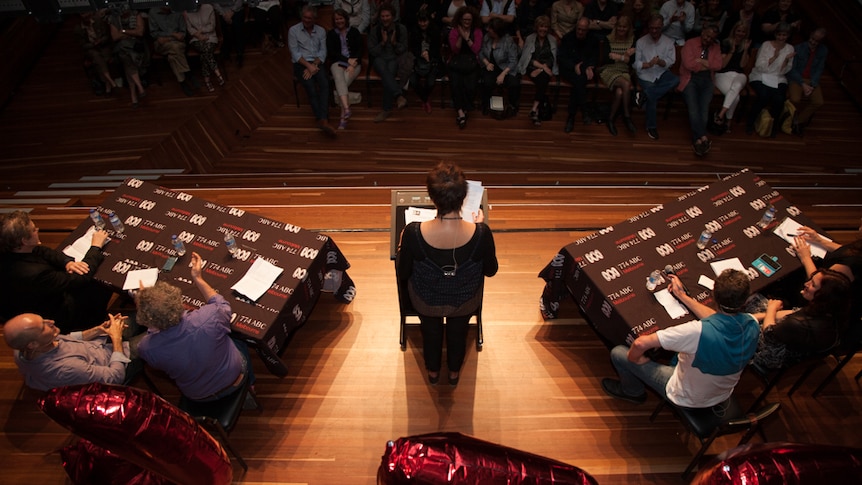 A crowd face a stage featuring a person on a lecturn flanked by two tables of three people.