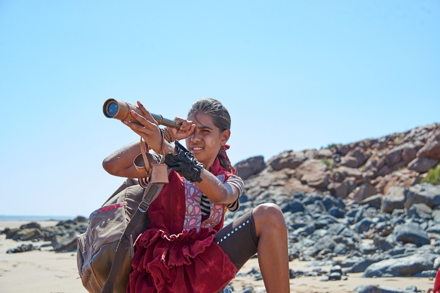 A young girl crouches near rocky beach landscape and holds brass handheld telescope to face on sunny clear blue sky day.