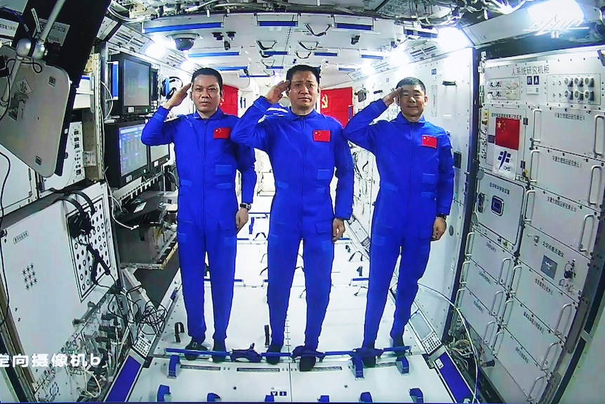 Three men in blue jumpsuits with Chinese flags on the front stand inside a space station, saluting to the camera. 