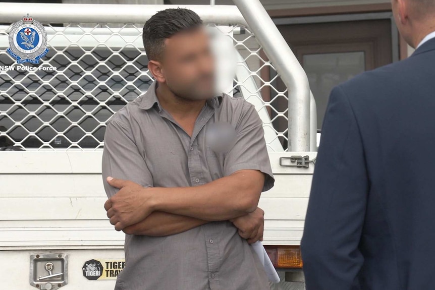 A man whose face has been blurred stands against a ute with his arms crossed and talks to detectives.