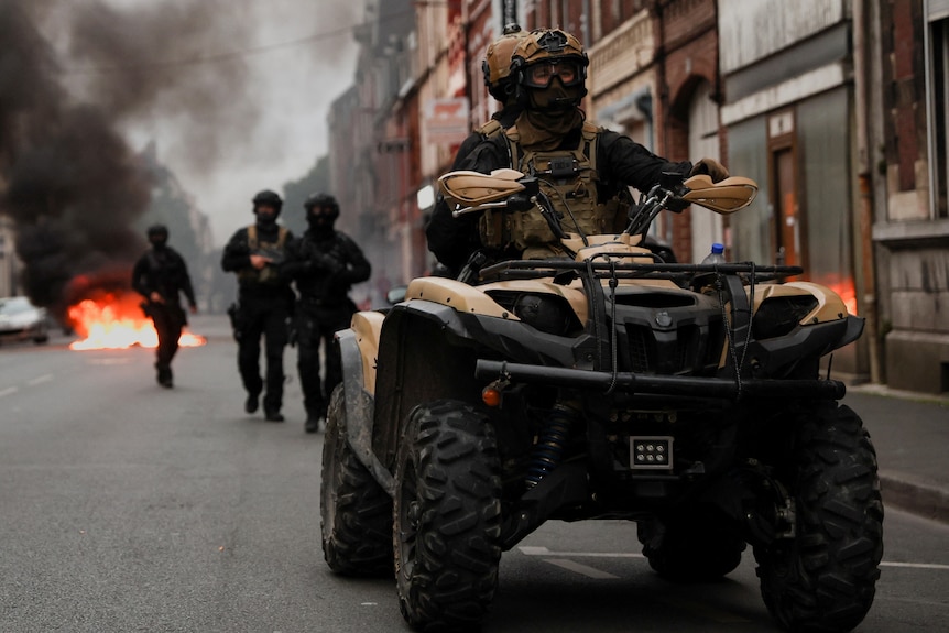 Two officers drive a quad bike on a street in Lille, France, behind them a smoking fire