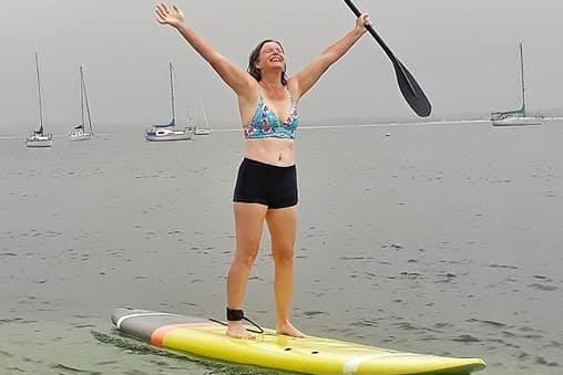 Nicola stand up paddle boarding in the rain, raising her arms in the air victoriously.