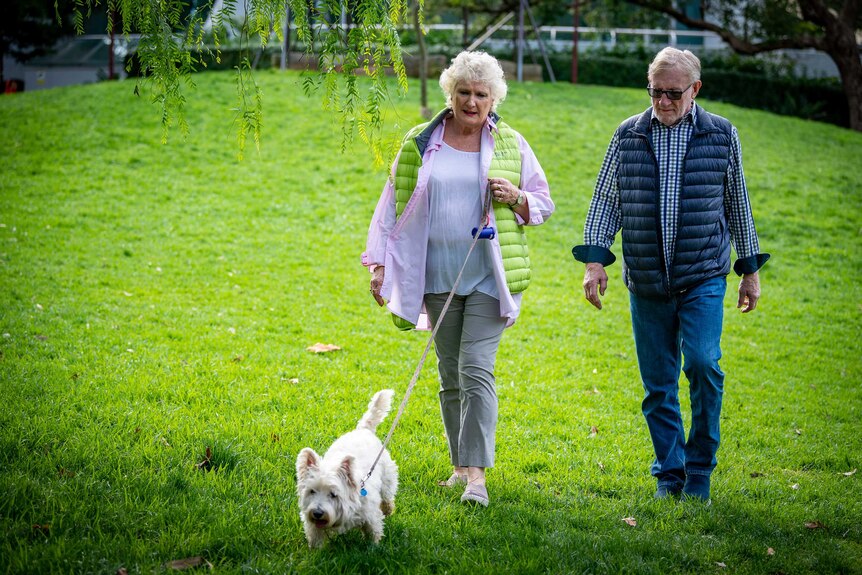 John and Bev Kable walk a small dog on a green lawn.