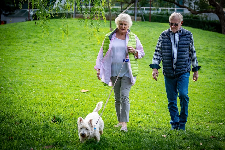 John and Bev Kable walk a small dog on a green lawn.