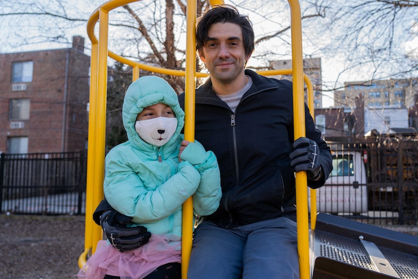 A man sits with a little girl in a green parker and face mask on a jungle gym