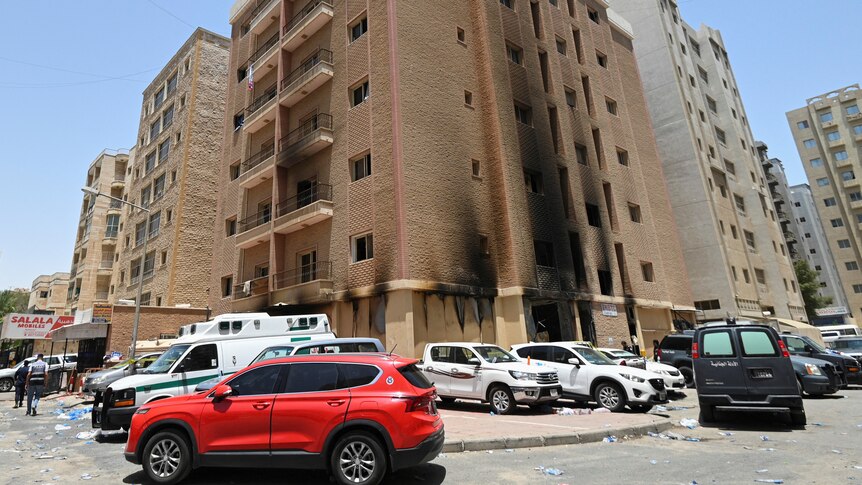 A brown six-storey building with some parts charred by a fire. There is a red car and other white cars parked around.