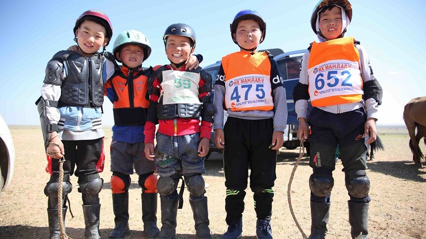 Five young Mongolian boys stand together in helmets and race bibs.