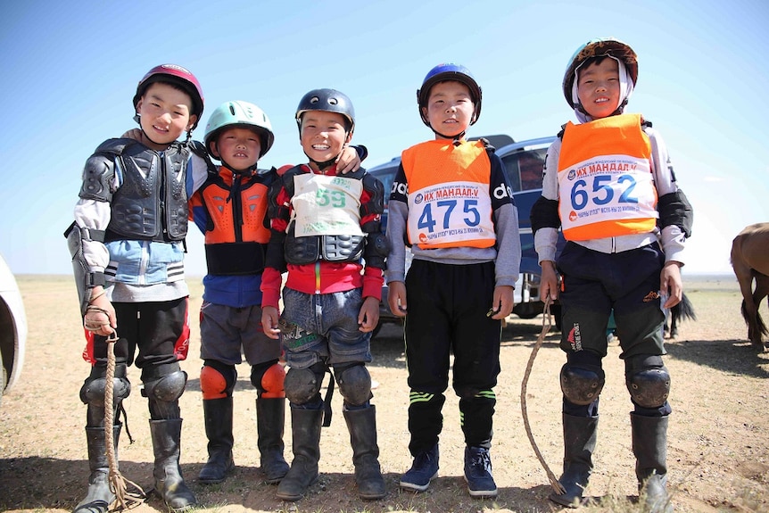 Five young Mongolian boys stand together in helmets and race bibs.