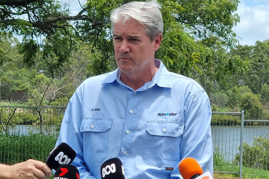 A mid shot of a man in a blue shirt standing in front of microphones