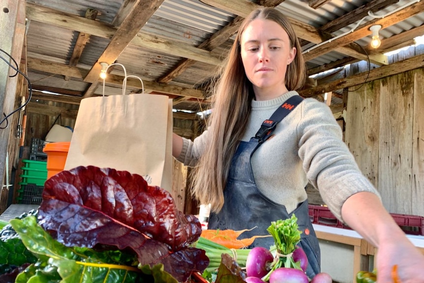 A woman in a black apron puts bunches of vegetables into a brown paper bag.