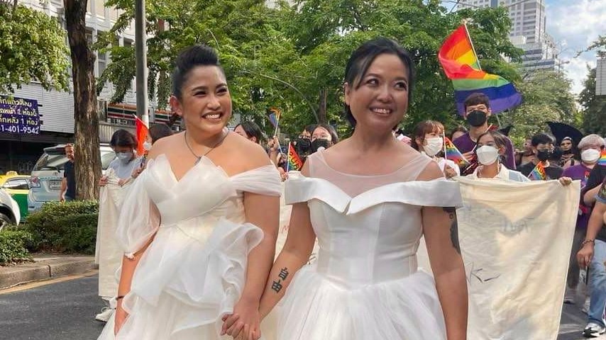 Two women in white puffy wedding dresses holding hands, smiling and marching in a pride festival with rainbow flag