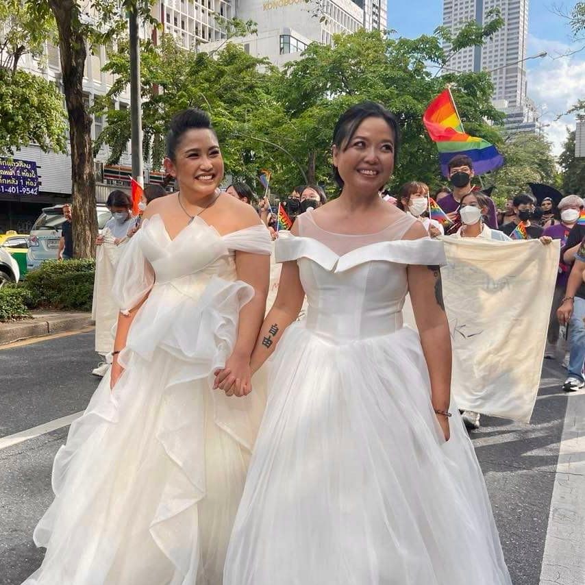 Two women in white puffy wedding dresses holding hands, smiling and marching in a pride festival with rainbow flag