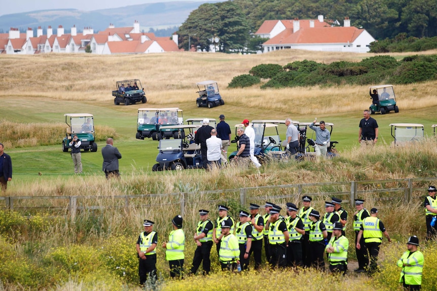 20 police in high-visibility uniforms stand in a clump behind a fence as a group of men stand near golf buggys