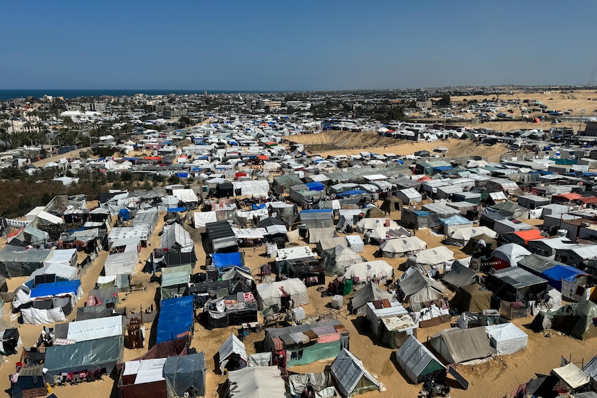 An aerial view of hundreds of tents set up on brown sand across a stretch of land with a blue sky in the distance.