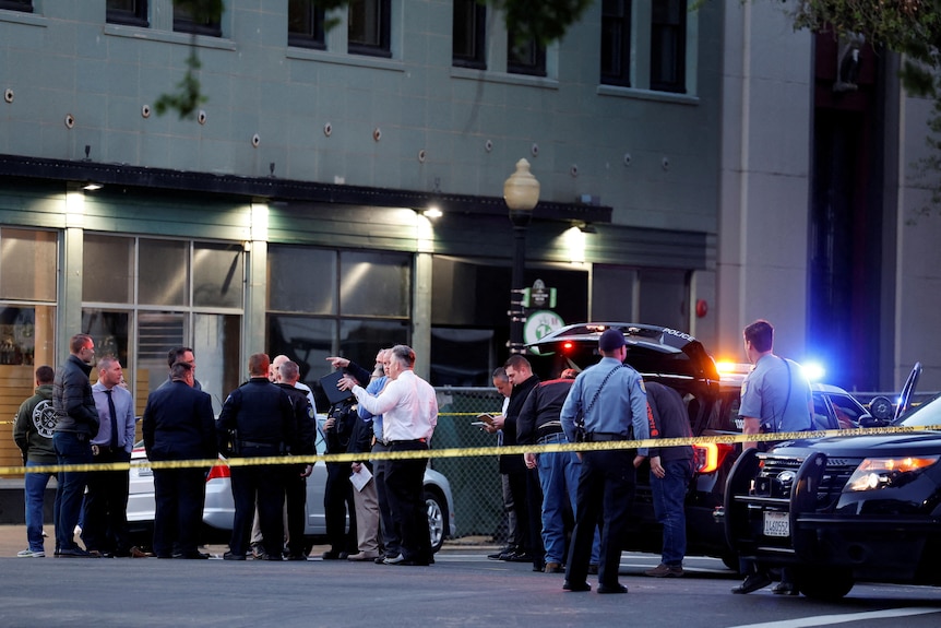 More than a dozen police and detectives, mostly men, stand and chat behind police tape, next to three vehicles.