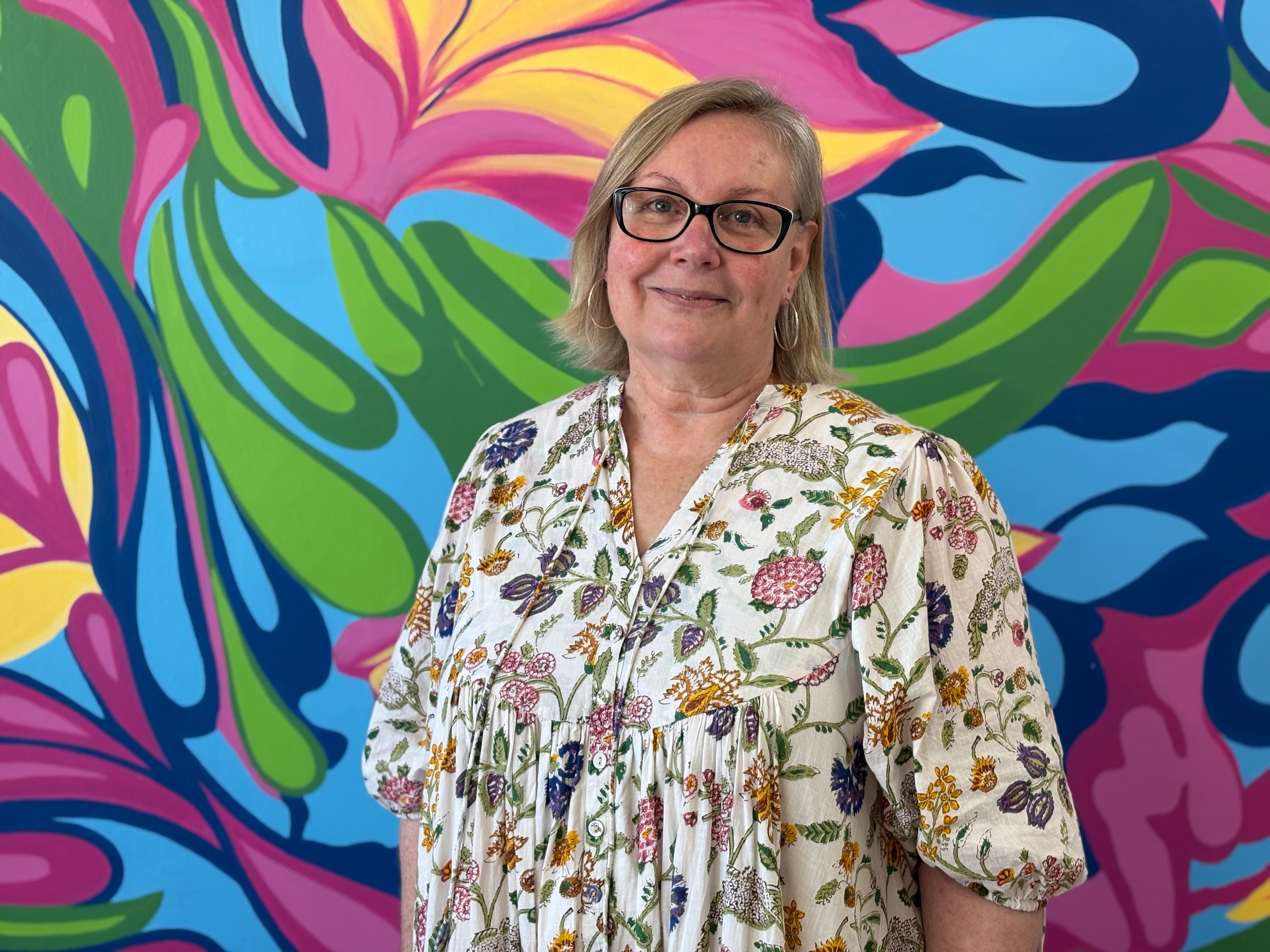 Μια γυναίκα με γυαλιά με σκούρο πλαίσιο και κοντά μαλλιά, φορώντας ένα φωτεινό λουλουδάτο φόρεμα στέκεται μπροστά από μια πολύχρωμη τοιχογραφία.