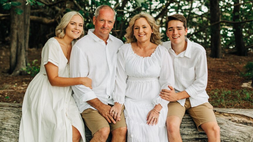 A family portrait under trees of a couple and their young adult children. All smiling and dressed in white.