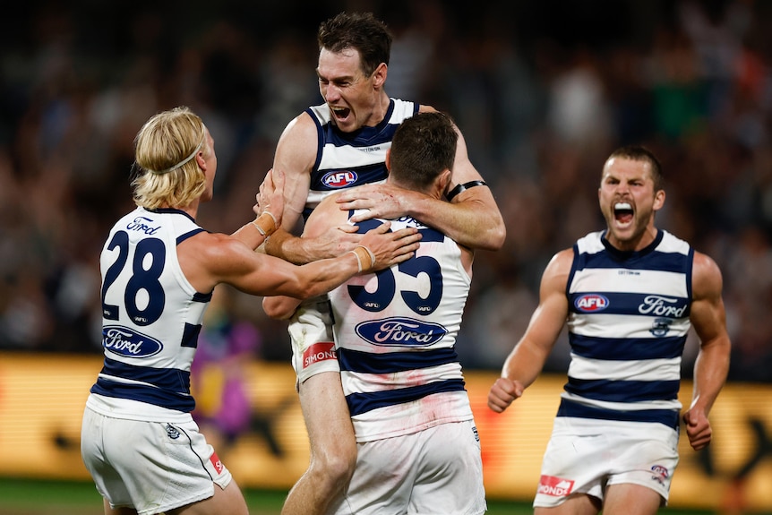A Geelong AFL captain lifts a teammate in the air as two other players celebrate next to them after a vital goal.