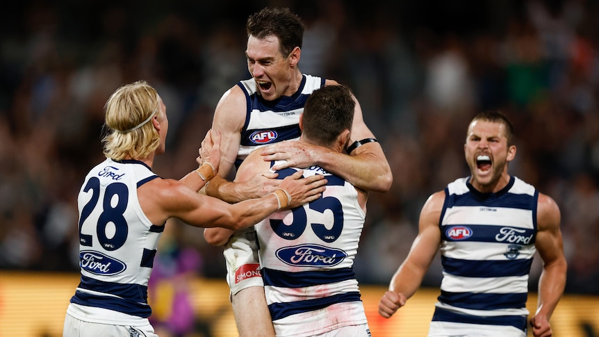 A Geelong AFL captain lifts a teammate in the air as two other players celebrate next to them after a vital goal.