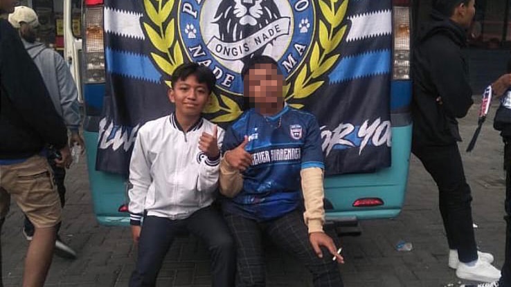 A teenage boy sitting next to an older boy in front of a Indonesian football flag.