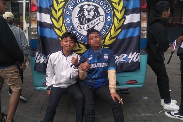 A teenage boy sitting next to an older boy in front of a Indonesian football flag.