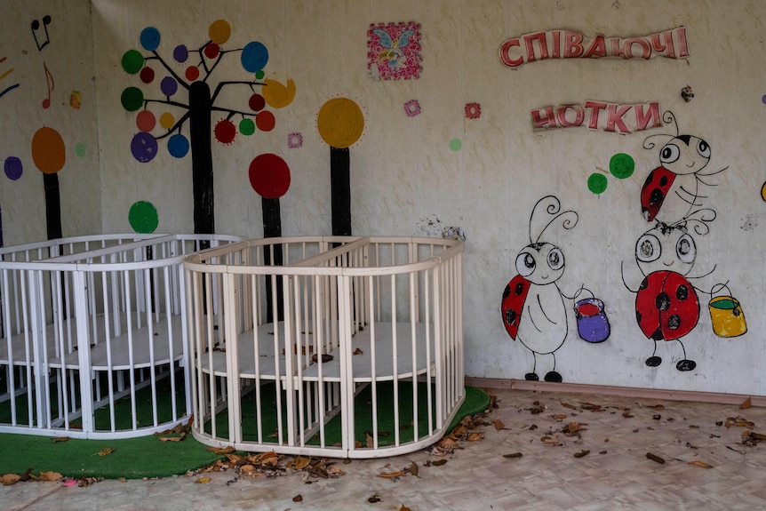 Two empty cots sit in a room that has leaves on the floor and bright art on the walls. 