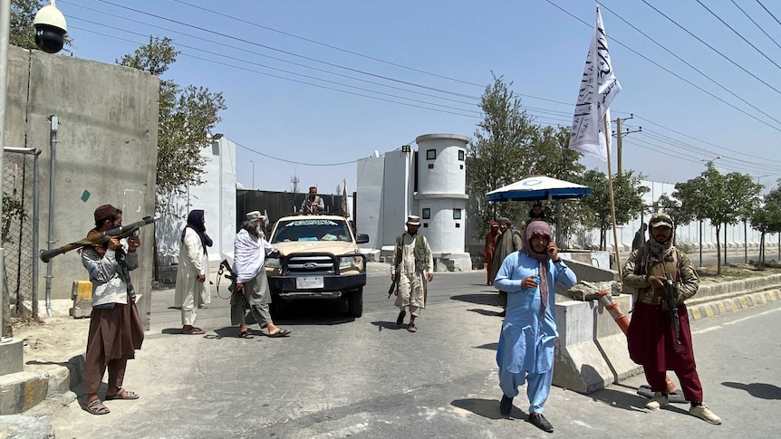 armed members of the taliban stand guard at an entrance gate in Kabul. taliban flag stands on right hand side.