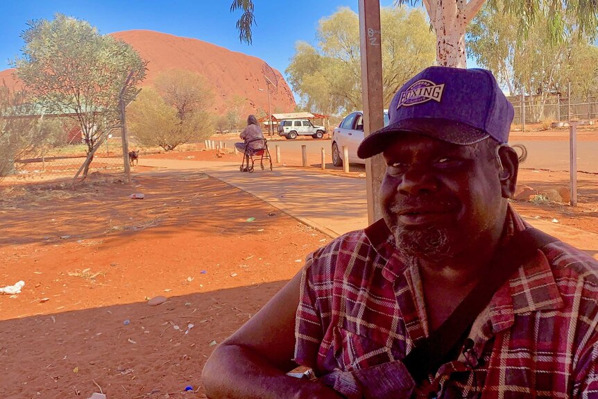 An Aboriginal man sitting in the shade with a cap on with Uluru in the background