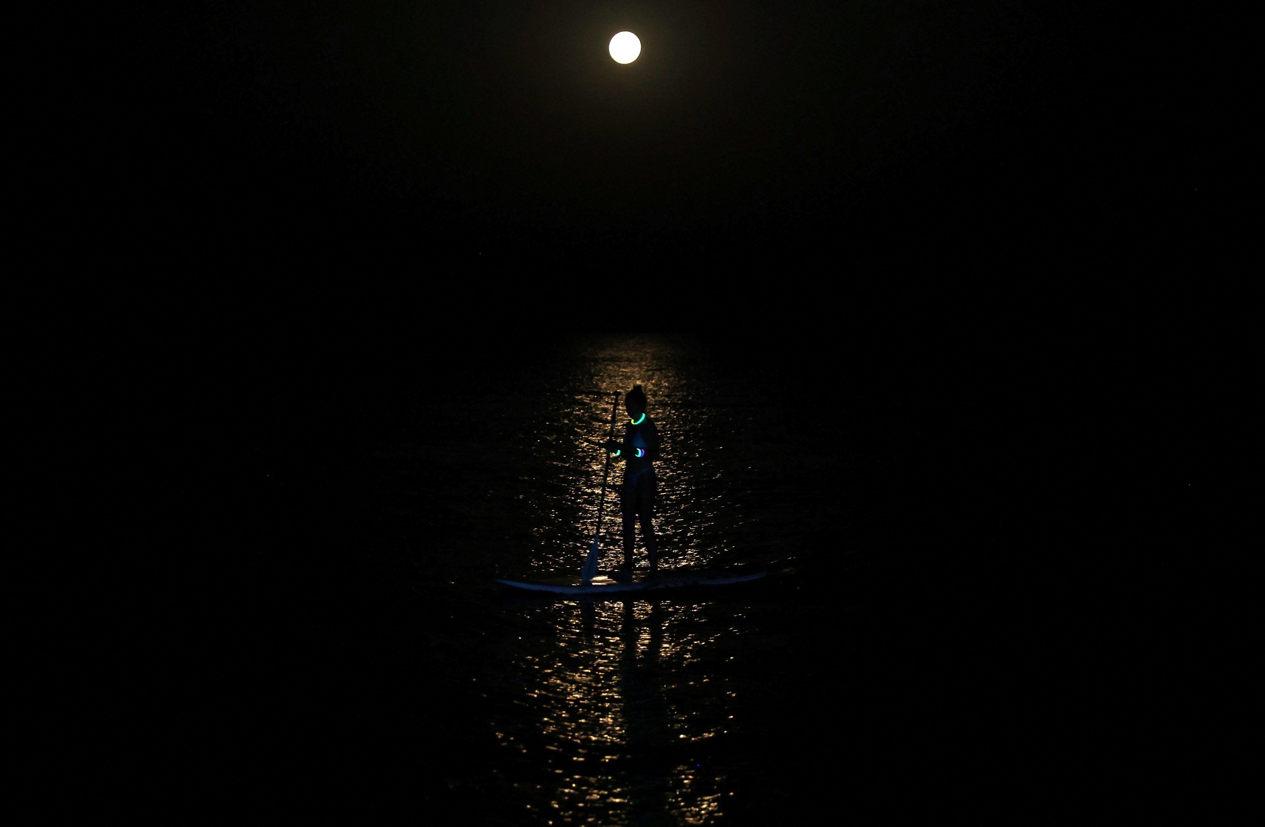 A person at night paddles a narrow board standing up,  a sliver of light illuminating them.